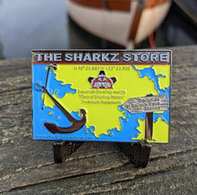 Rectangular geocoin sitting on a railing. Coin graphic is a map of esquimalt surrounded by water with 3D anchor and a sign that says 'dockside at sailor's cove marina' pointing to the location of the Sharkz Store. Banner at the top of the coin says The Sharkz Store. Colours are bright green for the land and bright blue for the water. 
