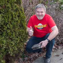 Man wearing a red Cachezinga T-Shirt holding a camo geocache doing a thumbs up