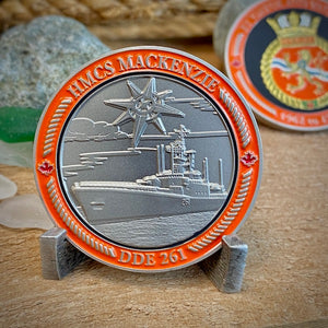 Back view of MacKenzie challenge coin in a display stand. The coin displays the Mackenzie Ship in the water with a compass star above it, all surrounded by a border with "HMCS MACKENZIE" at the top, and "DDE 261" at the bottom