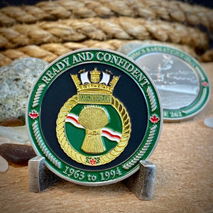 Front view of HMCS Saskatchewan challenge coin in a display stand. The coin displays the Saskatchewan Ship crest surrounded by a border with the phrase "Ready and Confident" at the top, and "1963 to 1994" at the bottom