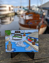Rectangular geocoin sitting on a railing. A marina with boats and a houseboat is visible in the background. image on the coin is the same view, with the addition of a swan, a seagull  with a french fry in it's beak and a seal. 