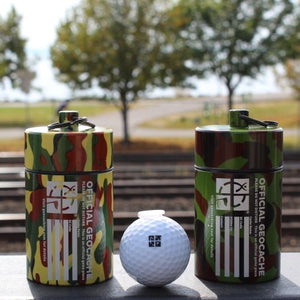 Cache tube containers in both variations next to a golf ball for size reference. The light camo is yellow, orange, and light green - where as the dark camo is dark green, light green, and brown.