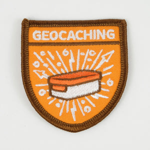 Shield shaped orange patch with a brown border.  It says "Geocaching" at the top and has a picture of a geocache under it. 